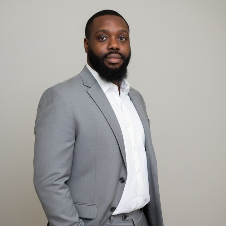 Man in a gray suit and white shirt stands confidently against a light background, showcasing a well-groomed beard.